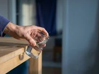 Mão segurando um copo de vidro na beira de uma mesa de madeira, prestes a cair