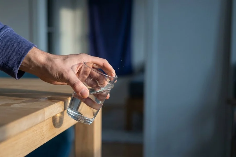 Mão segurando um copo de vidro na beira de uma mesa de madeira, prestes a cair